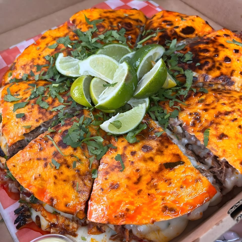 A large, sliced birria pizza from Tacos Mexiko, topped with cilantro and lime wedges.
