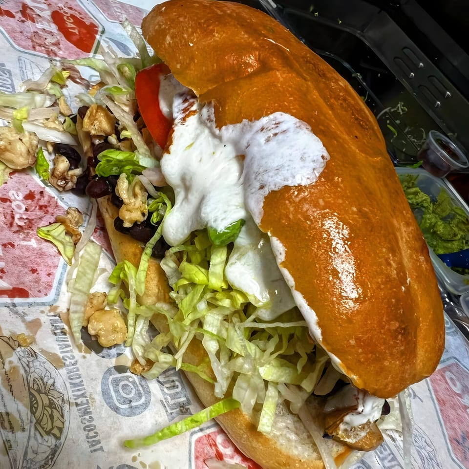 A large Tacos Mexiko Mexican torta, filled with meat, beans, lettuce, tomato, and crema.