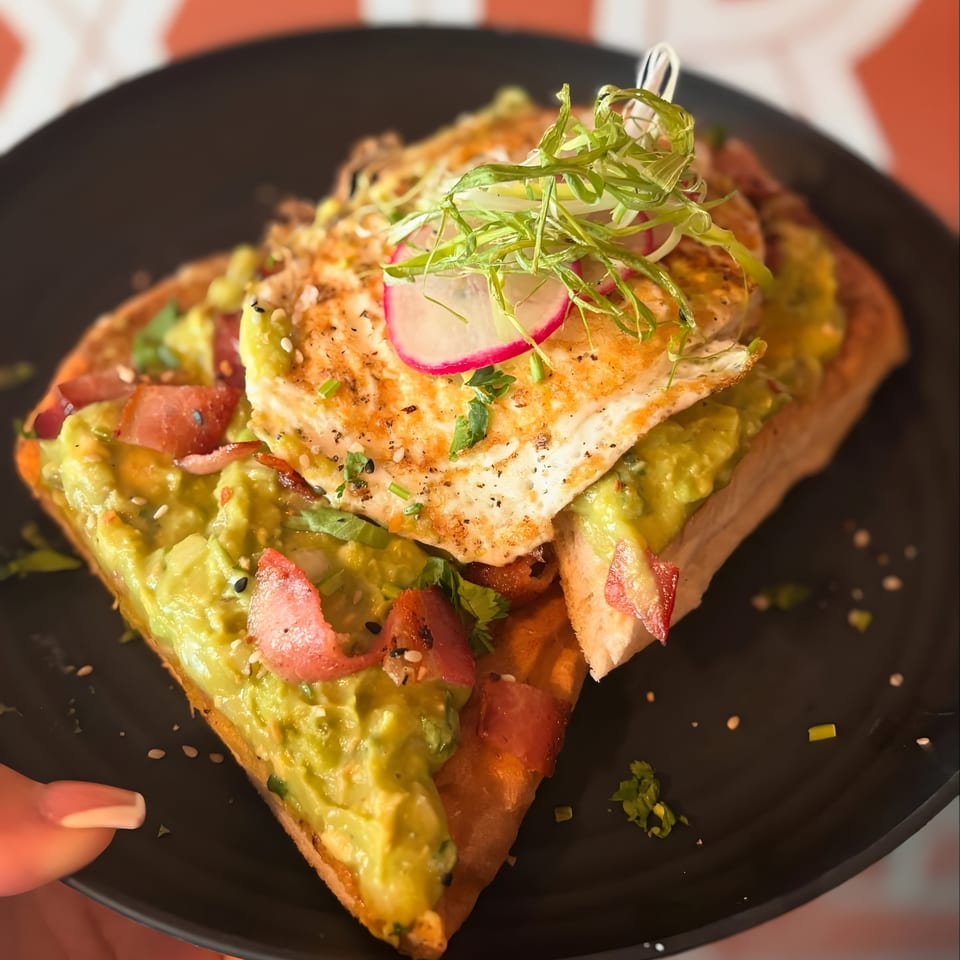 Avocado toast with bacon, a fried egg, radishes, and green onions on a black plate.