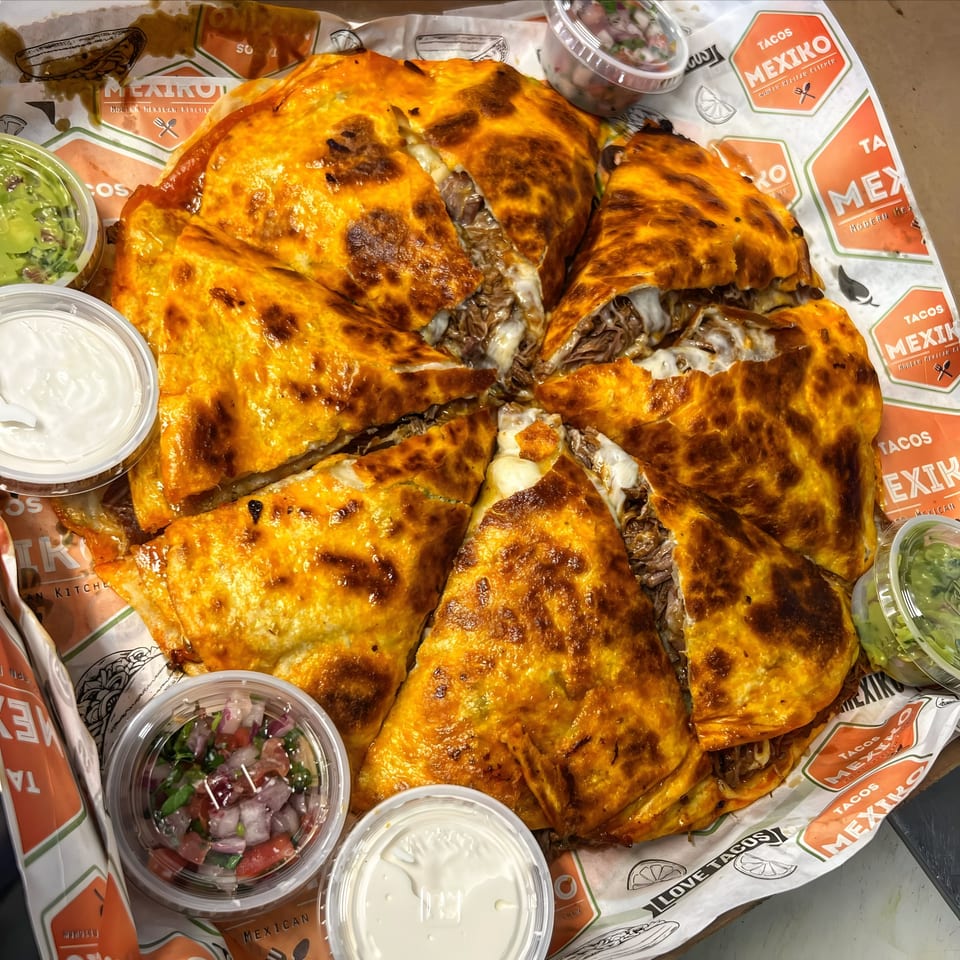 A large, sliced Tacos Mexiko quesadilla with meat and cheese, surrounded by dipping sauces.