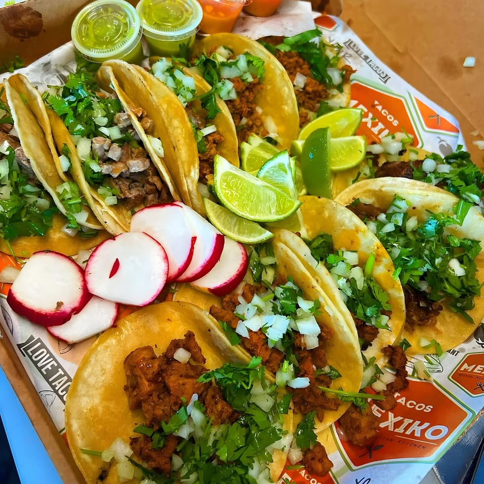 A box of Tacos Mexiko tacos with various fillings, cilantro, onions, lime wedges, and radish slices.